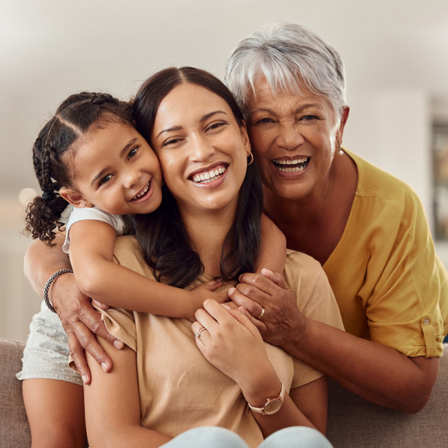 Three generations of smiling women hugging indoors, representing family dental care.