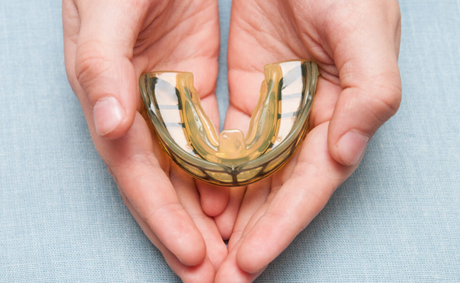 Hands holding a gold dental crown or veneer against light blue background.