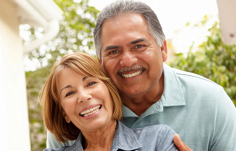 Smiling middle-aged couple posing together outdoors in casual clothing near greenery.
