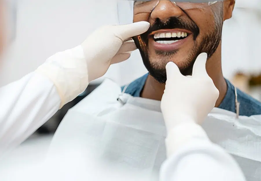 Male patient smiling during dental examination with dentist's hands checking teeth in clinical setting.