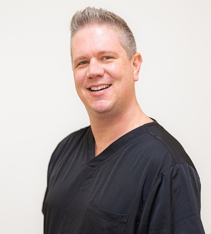 Professional headshot of male dentist smiling at camera wearing black scrubs against white background