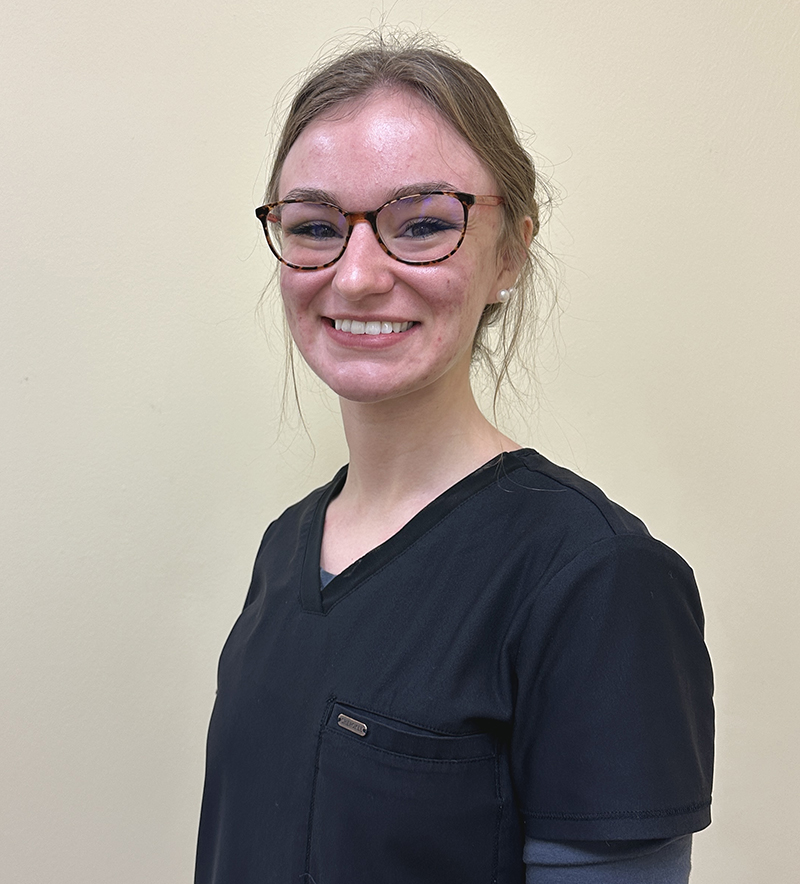Young woman wearing glasses and dark scrubs smiles at camera in clinical dental office setting.