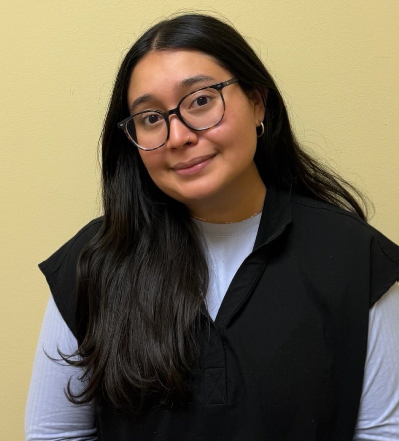 Young woman with glasses wearing black vest over light blue shirt against yellow background.